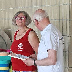 Zwei ältere Personen im Gespräch am Schwimmbadrand, eine Frau im roten Shirt mit Logo, ein Mann hält ein Klemmbrett.