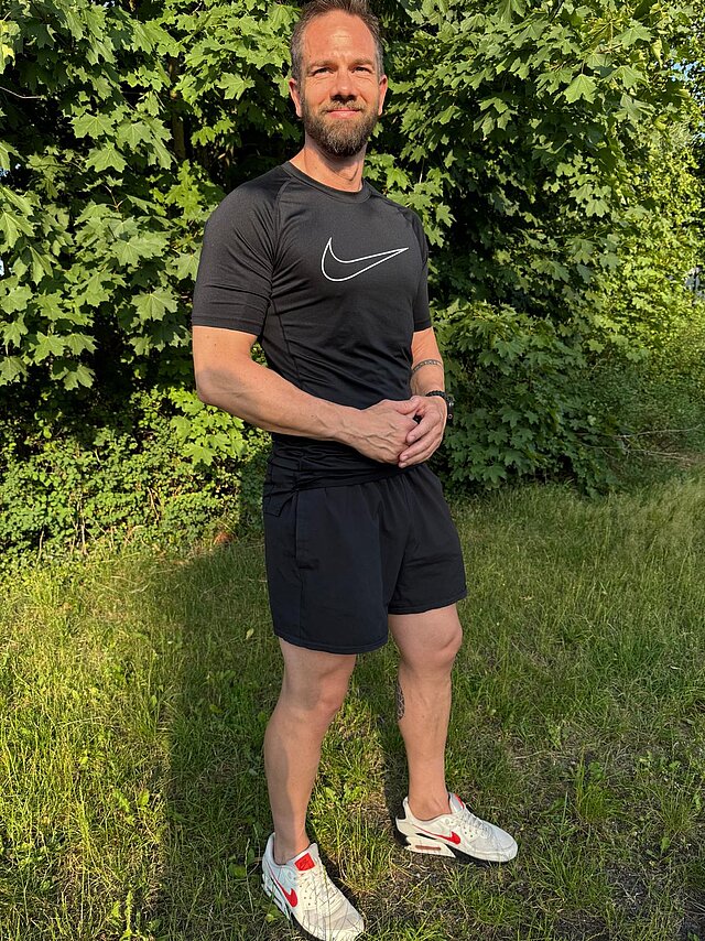 Tobias Breitenfellner Sportlich gekleideter Mann in schwarzem Shirt und Shorts steht draußen auf Gras vor dichtem Laub und lächelt.